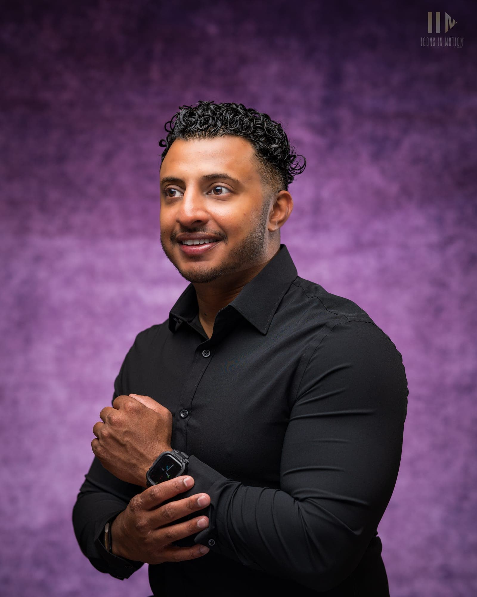 Studio headshot of a person with curly hair in a black shirt, arms folded, smiling against a purple backdrop