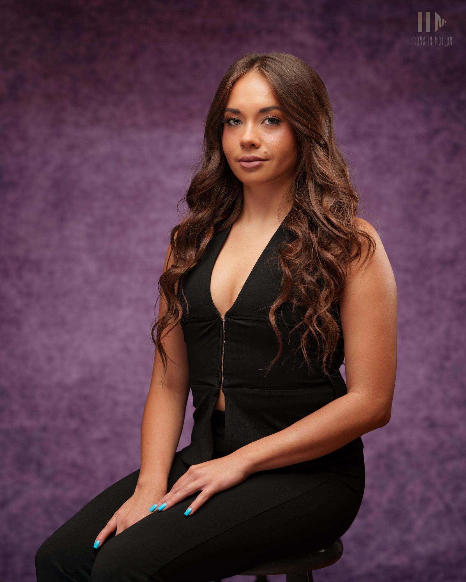 Full-length studio portrait of a person with long wavy hair seated on a stool