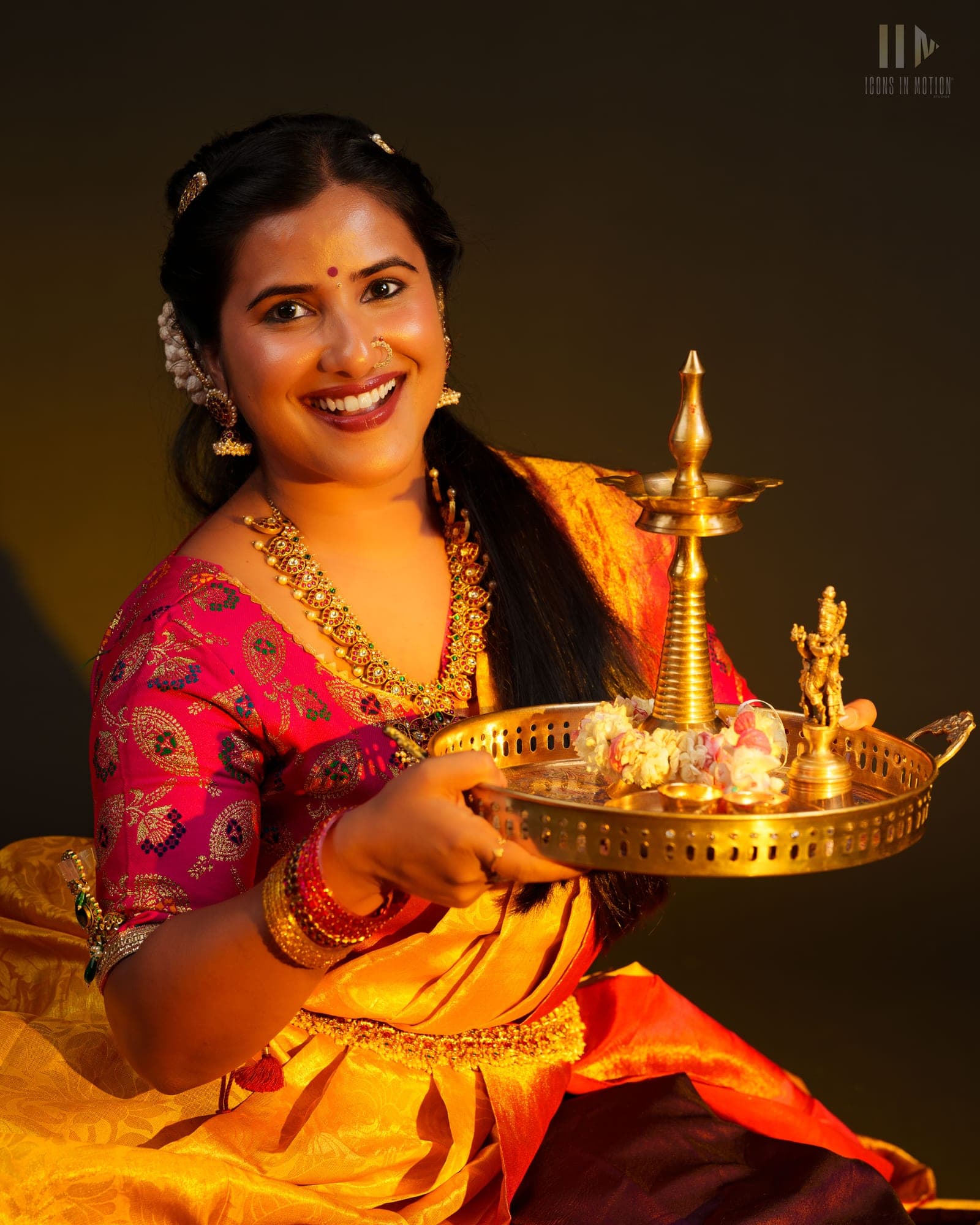 Dancer in a gold and pink saree holding a lit brass lamp tray