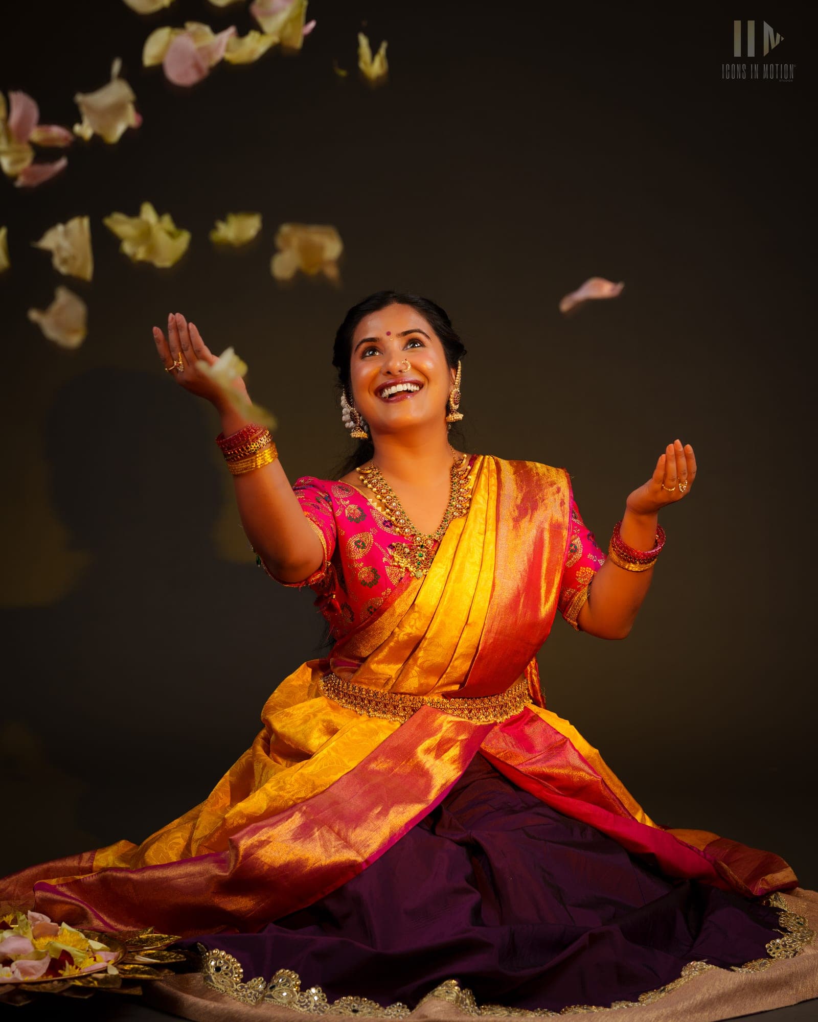 Dancer kneeling in a gold and pink silk saree as flower petals fall