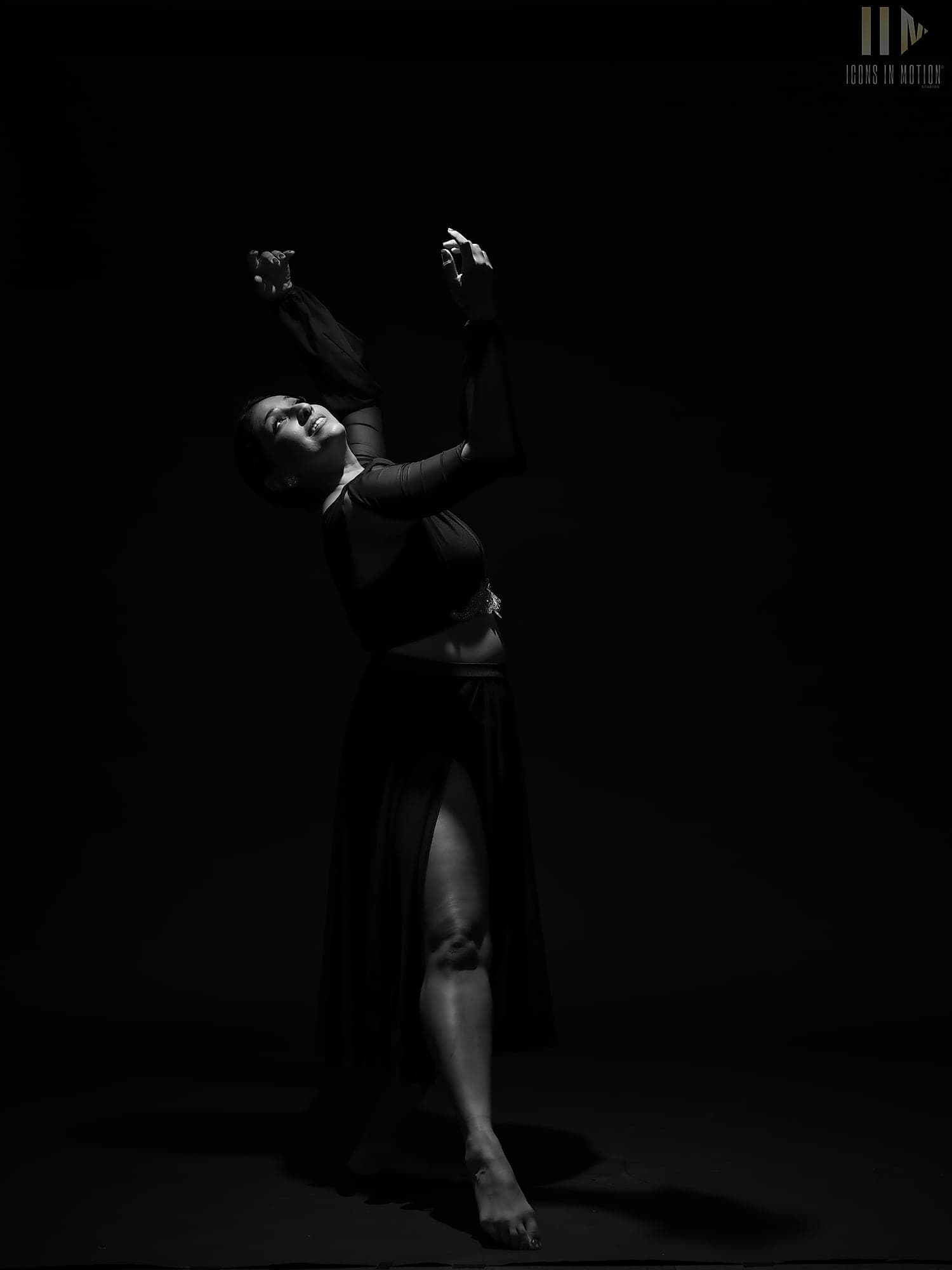 Dancer in black arching back with both hands raised, lit by a single light against darkness